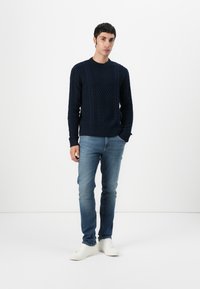 Marineblauwe gebreide trui met een textuur van kabelpatroon, gecombineerd met lichtblauwe jeans en witte sneakers, tegen een effen witte achtergrond.