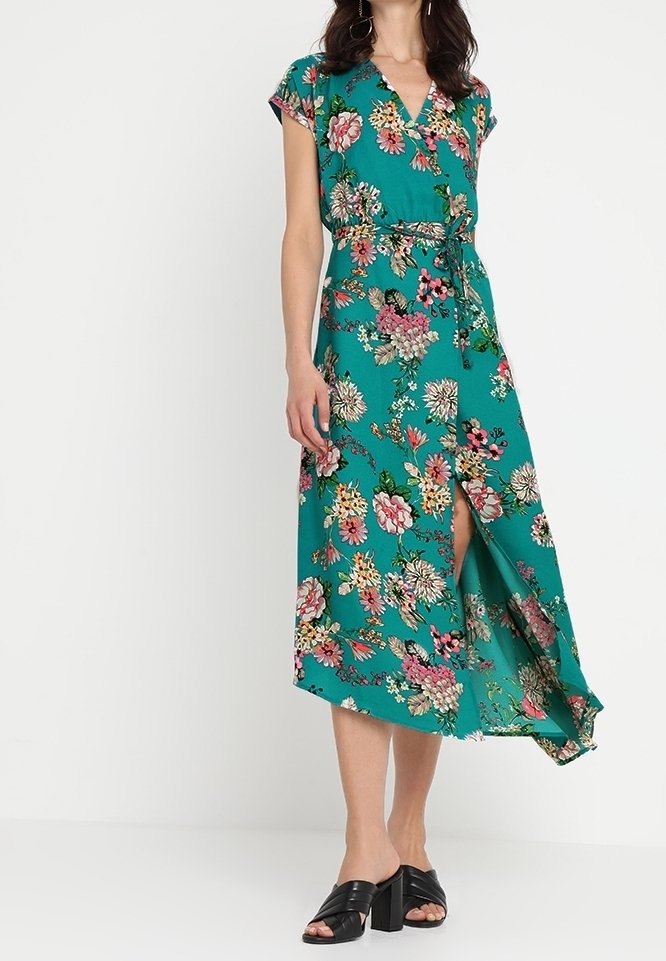 Femme portant une robe portefeuille à fleurs turquoise avec des manches courtes et des sandales à talons noires, se tenant contre un fond blanc uni.