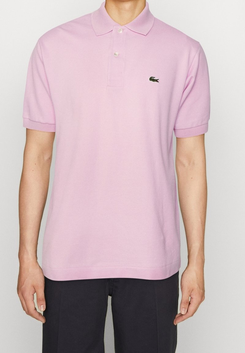 Polo en coton rose clair, avec un col à deux boutons et des manches courtes ; il présente un petit logo vert sur la poitrine gauche.