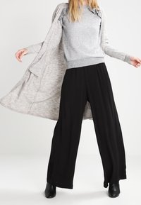 Hellgrauer langer Cardigan über einem strukturierten grauen Pullover, kombiniert mit lockeren schwarzen Hosen und schwarzen Stiefeletten.