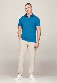 Jovem em pé, vestindo uma camisola polo azul, calças bege e ténis brancos, contra um fundo claro e liso.