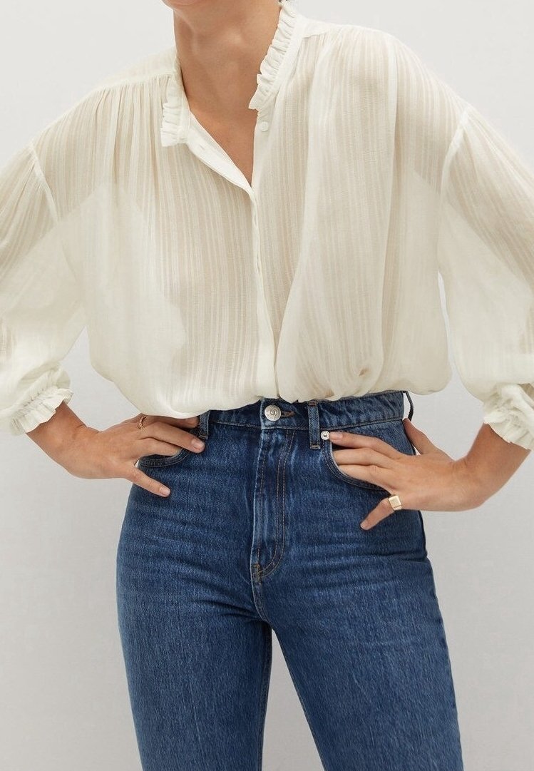 Blouse blanche à manches longues avec col et poignets à volants, associée à un jean bleu taille haute, mains posées sur les hanches, bague à la main droite.