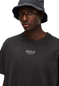 Sort bomulds t-shirt med rund hals og hvid "HALO" logo med tal på brystet. Sammen med en grå bucket hat.