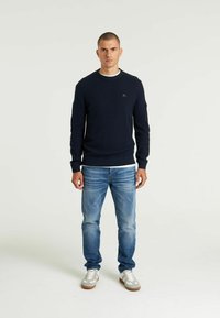 Dunkelblaues Strickpullover mit rundem Halsausschnitt und einem kleinen Logodetail auf der Brust, kombiniert mit hellblauen Jeans und grauen Sneakern.