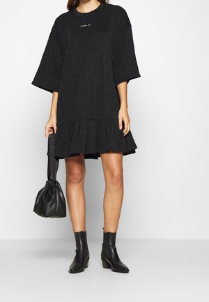 Frau trägt ein schwarzes, übergroßes Kleid mit ausgestellten Ärmeln und Saum, hält eine schwarze Leder-Clutch und trägt schwarze Stiefeletten.