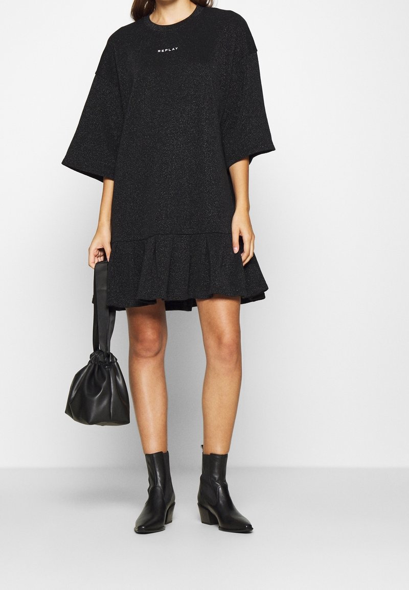 Femme portant une robe noire ample avec des manches et un ourlet évasés, tenant une pochette en cuir noir et des bottines noires.