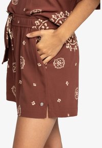 Pantaloni corti a motivo marrone con una vita regolabile tramite cordoncino, tasche laterali e dettagli in metallo argentato. Presentano disegni floreali e simili a mandala.