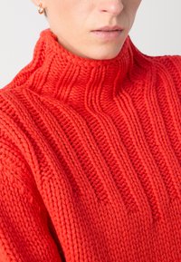 Roter gestrickter Rollkragenpullover mit strukturierten Rippmustern und grobem Gewebe, das enge und lockere Strickabschnitte zeigt.