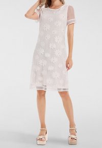 Femme portant une robe blanche en dentelle longueur genou avec des motifs floraux et des manches courtes, associée à des sandales compensées beiges.