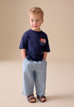 Niño pequeño con cabello rubio corto que lleva una camiseta azul marino con un gráfico de un camión de bomberos, pantalones azul claro y sandalias oscuras, de pie con las manos en los bolsillos.