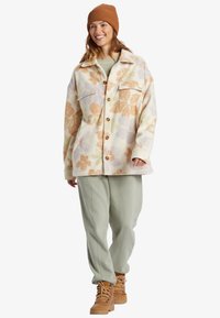 Fleece jacket with floral pattern in peach and pastel colors, features two chest pockets and button closure, paired with light green pants and boots.