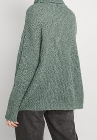 Zielony sweter z dzianiny z wysokim kołnierzem, w oversize'owym kroju, o fakturowanej tkaninie i opuszczonych ramionach. Prążkowane mankiety z luźną sylwetką.