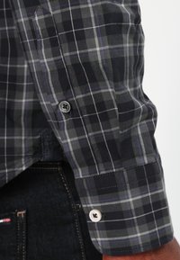 Camicetta a maniche lunghe a quadri neri e grigi con polsini e bottoni, indossata sopra dei jeans in denim scuro su un braccio di una persona.