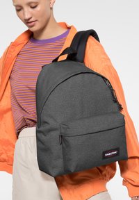 Mochila gris con un acabado de tela suave, que presenta un bolsillo frontal y una correa acolchada, exhibida con un modelo en atuendo casual.