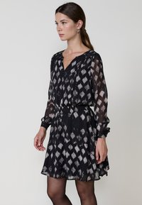Schwarz-weiß gemustertes Kleid mit V-Ausschnitt, langen Ärmeln und elastischem Bund. Hergestellt aus transparenter Stoff, mit einem fließenden Design und subtiler Sheen.