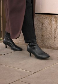 Black leather ankle boots with a pointed toe and thin stiletto heel, featuring a smooth, fitted design. Worn with dark jeans and a long coat.