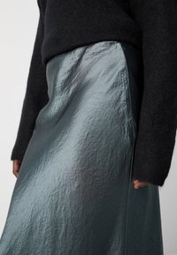 Metalizuota teal sijonas pasižymi tekstūruotu paviršiumi ir elegantišku dizainu, derinamas su juodu megztiniu. Matoma satino paviršius ir šoninė kišenė.