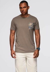 Hnědý bavlněný tričko s vzorovanou kapsou v zelené, žluté a modré barvě. Krátké rukávy, kulatý výstřih, spárované se světlými kalhotami.