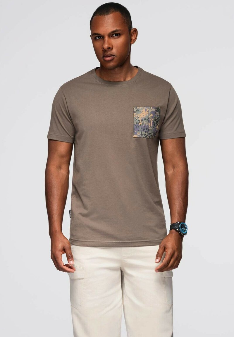 Hnědý bavlněný tričko s vzorovanou kapsou v zelené, žluté a modré barvě. Krátké rukávy, kulatý výstřih, spárované se světlými kalhotami.