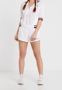 Mono corto estilo polo blanco con mangas cortas, presenta rayas rojas y azul marino, cintura elástica y detalle de logo en el dobladillo. Combinado con zapatillas blancas.