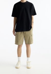 Camiseta negra de gran tamaño, pantalones cortos de cargo color caqui con bolsillos laterales, zapatillas blancas y calcetines blancos de canalé. Diseño simple, material de algodón.