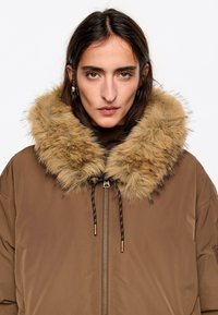 Chaqueta acolchada marrón con cuello de piel beige peluda, con cierre frontal de cremallera y cordón ajustable. Textura lisa con detalles en herrajes dorados.