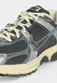 Tummanharmaat ja hopeiset Nike Vomero 5 -tennarit, joissa kermanväriset nauhat ja pehmustettu pohja, lähipotretissa näkyy teksturoidut ja kiiltävät materiaalit.