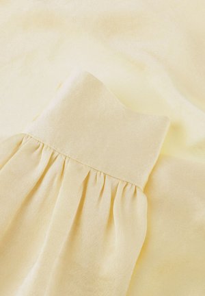 Zacht lichtgeel stof met een geplooid detail, waardoor zachte plooien en textuur ontstaan op een gladde achtergrond.