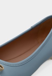 Modrá kožená balerína s bledě hnědou vnitřní podšívkou a béžovým detailem smyčky na patě, zobrazena zblízka na bílém pozadí.