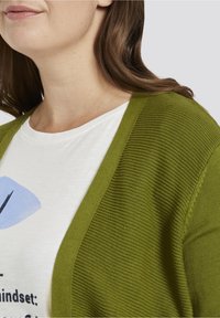 Cárdigan verde de canalé con abertura en la parte delantera, llevado sobre una camiseta blanca con texto y gráficos en azul y negro. Material de punto texturizado.