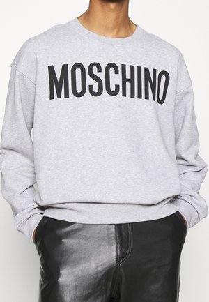 Persoon draagt een grijze trui met 'MOSCHINO' in zwarte letters en zwarte leren broek, met handen in de zakken, tegen een effen achtergrond.