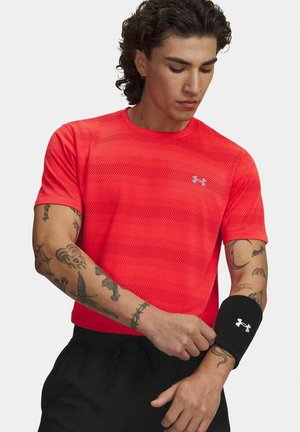 Man met krullend haar die een rood Under Armour-shirt draagt, past een zwarte polsband aan en toont meerdere tatoeages op beide armen.