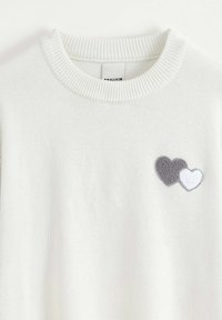 Pull en tricot blanc avec un col côtelé et un patch en forme de cœur gris et blanc sur le côté gauche. Texture douce, design décontracté.