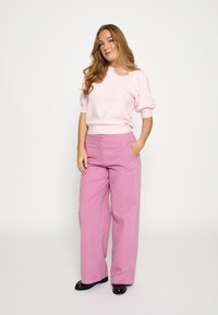 Lichtroze trui met korte mouwen en lichtroze wijde broek met hoge taille, voorzien van zijzakken en een gladde stoftextuur. Zwarte schoenen.