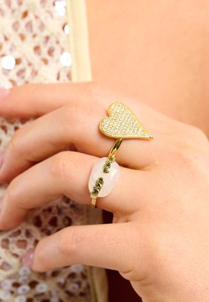 Bague en forme de cœur en or avec des cristaux clairs, associée à une bague à pierre ovale avec des accents verts, portée sur une main contre un fond texturé.