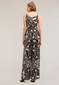 Svartvitt mönstrad jumpsuit med vida ben, V-back design och tunna axelband. Bärs med gröna högklackade skor, vilket visar en flödande textur.