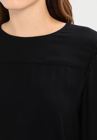 Schwarzes Oberteil mit rundem Ausschnitt und gerafftem Schulterdetail, aus weichem Material gefertigt, mit dezenten Nähten entlang der Brust.