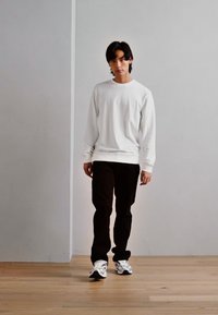 Ung mand går indendørs iklædt en hvid langærmet skjorte, sorte bukser og hvide sneakers på et trægulv med en ensfarvet væg i baggrunden.