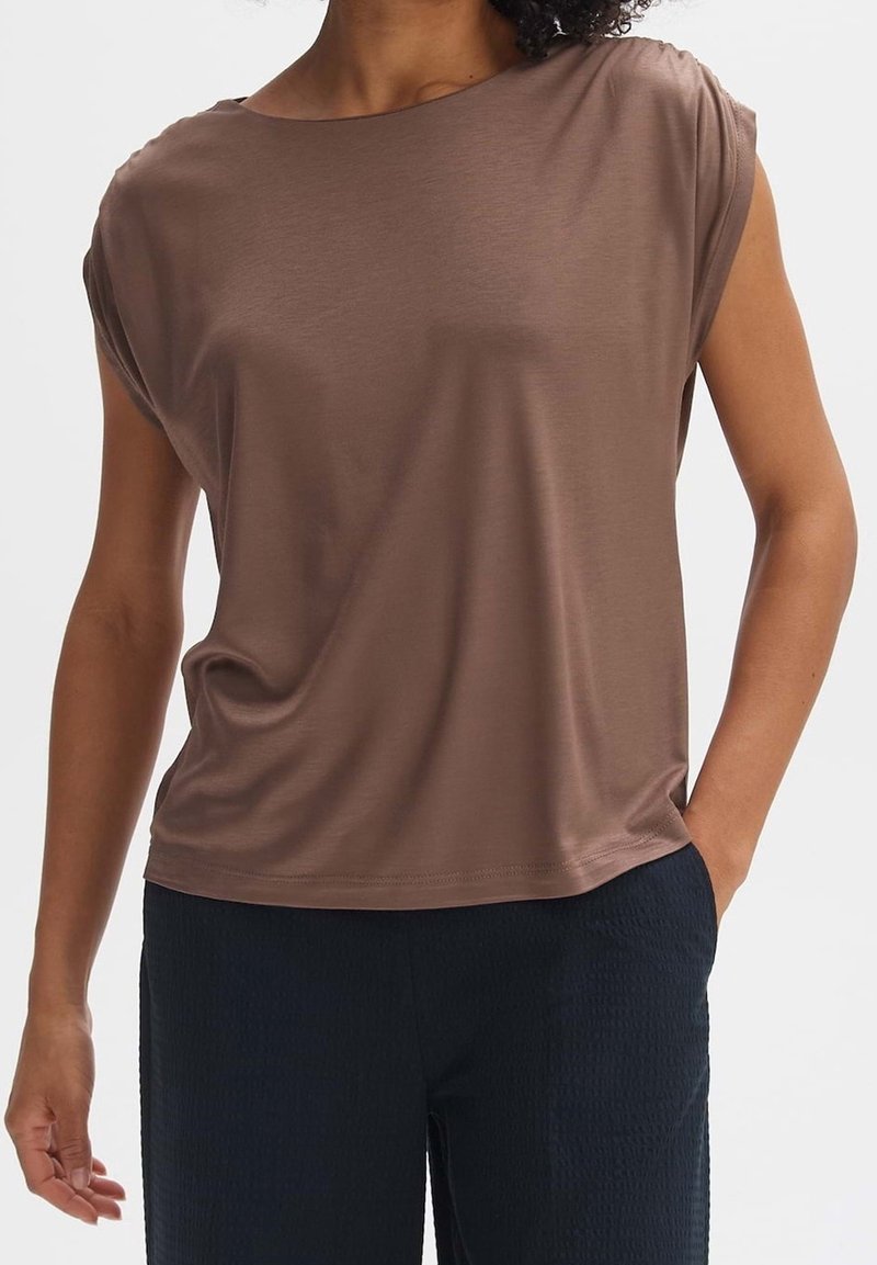 Femme portant un haut marron ample à manches courtes et un pantalon noir, une main dans la poche, sur un fond uni.