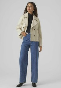 Beige dubbelknäppt kappa med stora slag över en svart topp, i kombination med högmidjade, vida blå jeans och svarta klackstövlar.