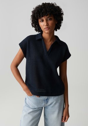 Frau mit lockigem Haar, die ein schwarzes Strickoberteil mit kurzen Ärmeln und hellblaue Jeans trägt, steht mit einer Hand in der Tasche vor einem neutralen Hintergrund.