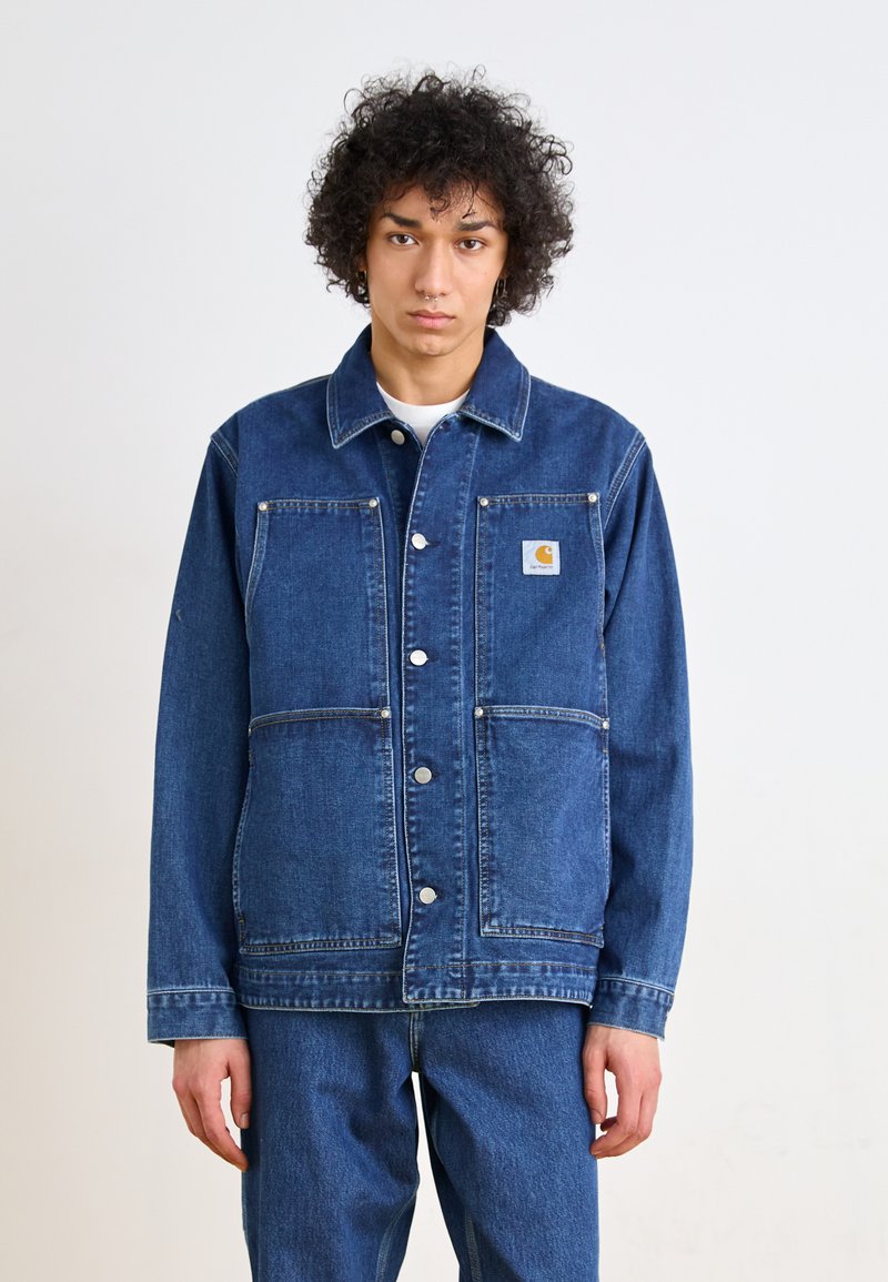 Jachetă din denim de culoare albastru închis, cu închidere cu nasturi, două buzunare pe piept și cusături contrastante. Detaliază o etichetă cu logo pe buzunarul din stânga.