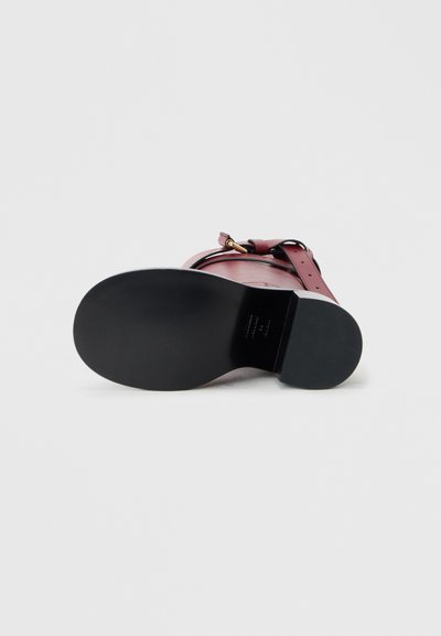 Burgundy leather sandal with a black rubber sole, featuring an adjustable strap and gold hardware accents. Bottom view.