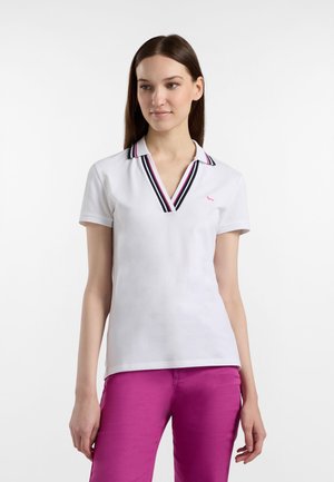 Nainen, joka pukeutuu valkoiseen lyhythihaiseen poolopaitaan, jossa on musta ja vaaleanpunainen raidallinen kaulus, yhdistettynä kirkkaan vaaleanpunaisiin housuihin, seisoo yksiväristä valkoista taustaa vasten.