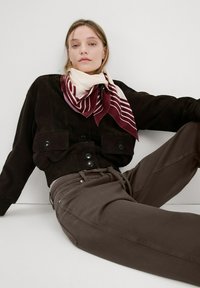 Mujer con chaqueta negra, pantalones marrones y bufanda a rayas burdeos y blancas, sentada con una pierna doblada sobre un fondo blanco.