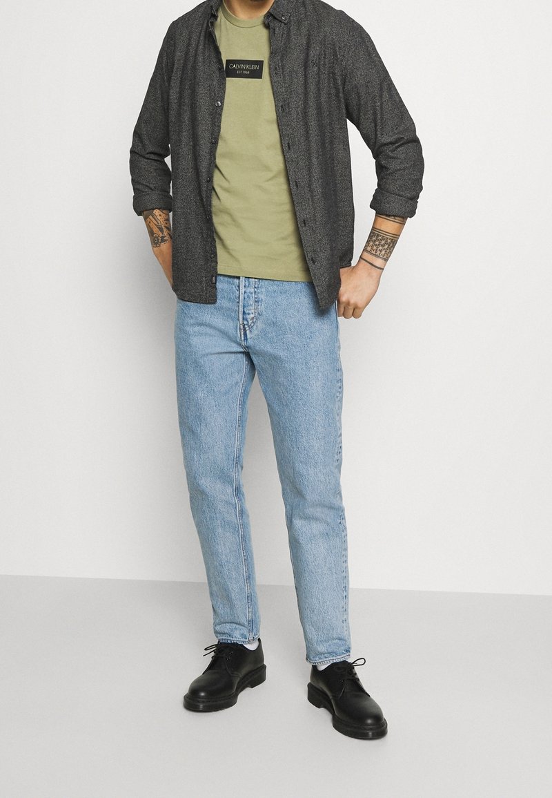 Homme portant une chemise grise boutonnée par-dessus un t-shirt olive Calvin Klein, un jean bleu clair, des chaussures noires et des tatouages visibles sur les deux avant-bras.