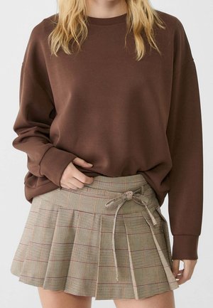 Osoba ubrana w brązowy sweter z długim rękawem oraz beżową plisowaną spódnicę w kratę z kokardą zawiązaną z boku.