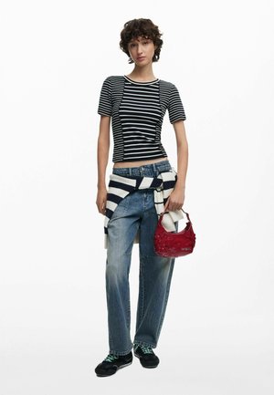 Jeune femme portant un haut court rayé noir et blanc, un jean bleu ample, des baskets noires, un pull rayé noué autour de la taille, tenant un sac à main rouge.