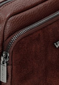 Bruine leren en suede tas met een gestructureerd oppervlak, zilverkleurige hardware met een ritssluiting en een logo-tag bevestigd.