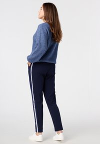 Marine joggers met witte zijkanten, gecombineerd met een getextureerde blauwe gebreide trui. Het model staat zijwaarts, met de handen in de zakken.